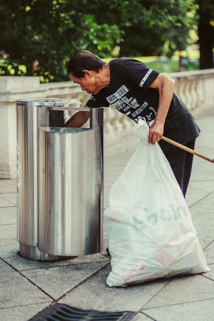 A woman with short dark hair, wearing a black T-shirt with white graphic print on the front, is seen on a paved outdoor area, bending slightly forward to dispose of trash into one of two cylindrical s